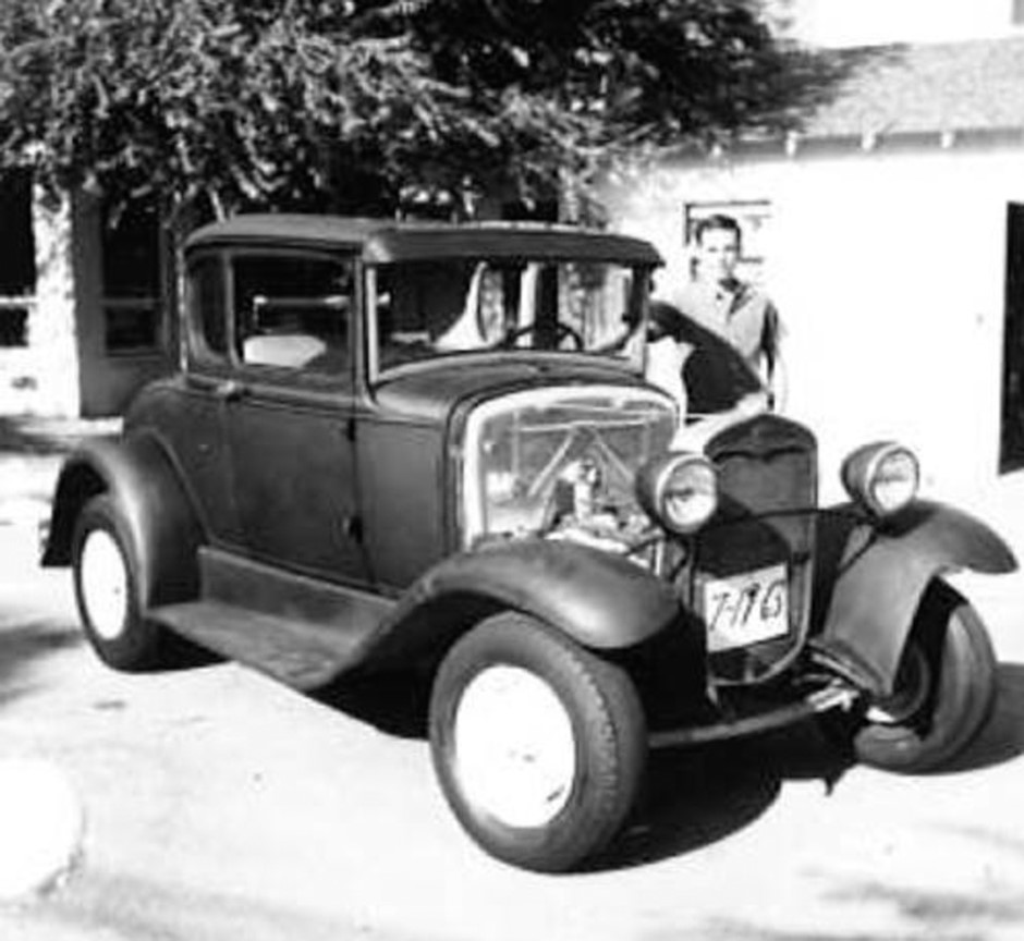 A black-and-white photo of Tony as a young man, standing outdoors beside a car.