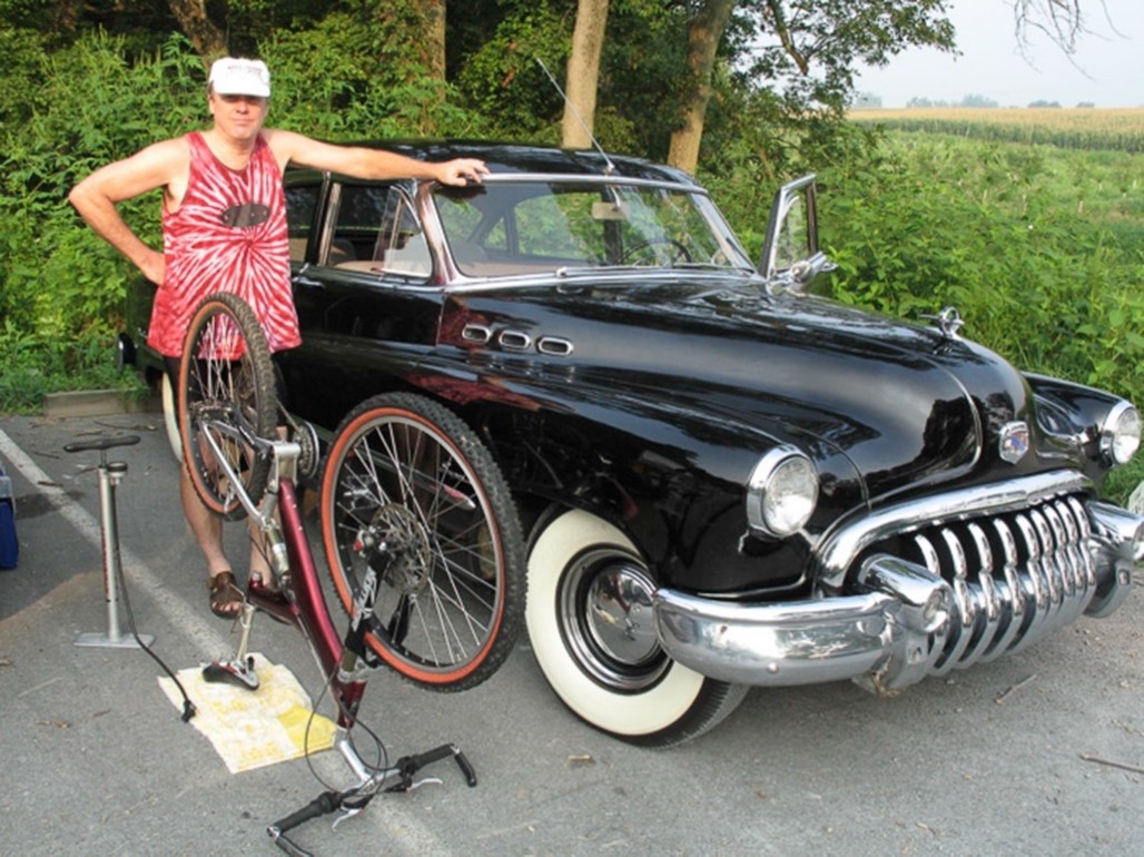 Tony standing outside by a black car with his arm resting along the hood. Nearby are a bike pump and a bicycle that's propped with its wheels up.